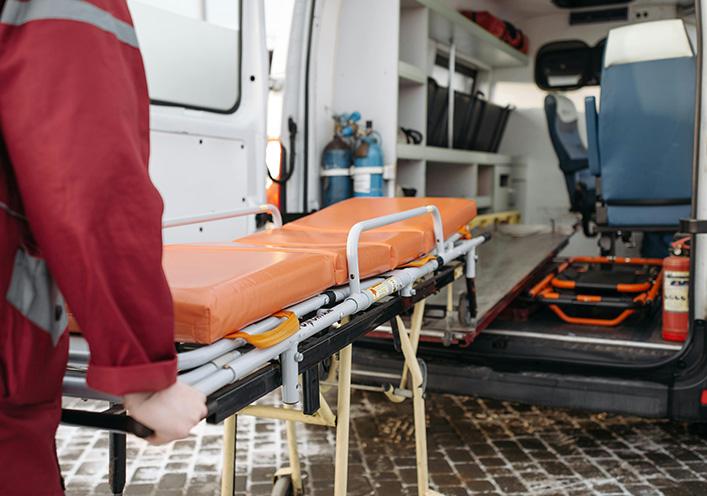 An emergency medical technician pushes an empty orange stretcher towards the open back of an ambulance.